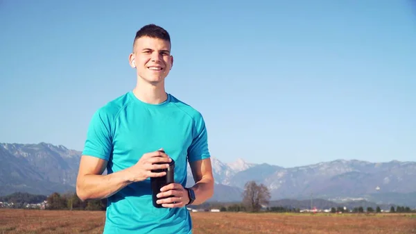 fit young man preparing protein shake and smiling in a camera with mountains the back