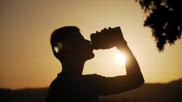 Risoluzione 4k di una sagoma di giovane uomo in forma che beve acqua con il sole dietro di essa al tramonto — Video Stock