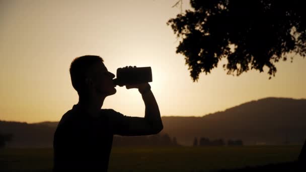 4k Auflösung einer Silhouette eines fitten jungen Mannes, der Wasser trinkt, mit Sonne im Hintergrund bei Sonnenuntergang — Stockvideo