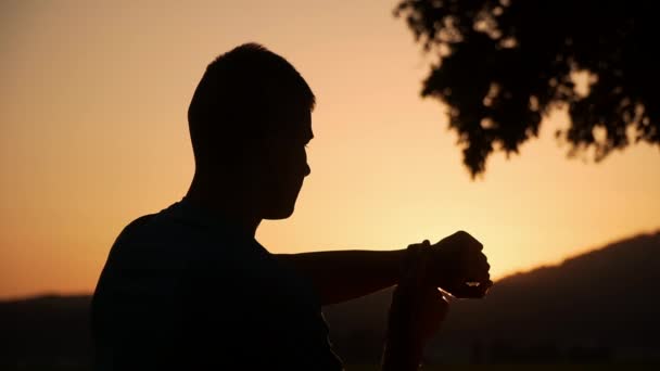 Sillhouette ενός άνδρα αγγίζοντας και ελέγχοντας το smartwatch στο όμορφο ηλιοβασίλεμα στο παρασκήνιο — Αρχείο Βίντεο