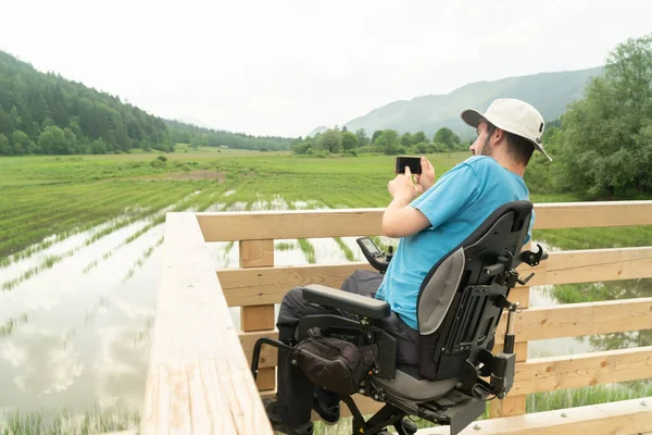 Hombre en silla de ruedas eléctrica con cámara de teléfono inteligente cerca del lago en la naturaleza —  Fotos de Stock