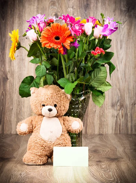 Um urso de pelúcia e flores brilhantes no vaso em fundo de madeira — Fotografia de Stock