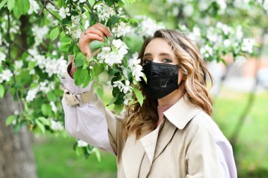 Arka planda maskeli bir kadın elma ağacı açıyor. Kavram ve sembol COVID-19 ve pandemik koronavirüsü karantinaya alır. 2020 baharında çiçek açan bahçede virüse ve alerjiye karşı koruma
