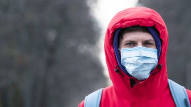 Yakın plan bir portre. Koruyucu, tek kullanımlık tıbbi maske takan genç Avrupalı bir adam dışarıda yürüyor. Yeni koronavirüs (Covid-19). Salgın veya salgın sırasında sağlık hizmeti kavramı