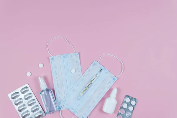 Protective medicine mask, antiseptic thermometer and pills on pink background, flat lay. Concept of doctor table and health care. Top view, copy space