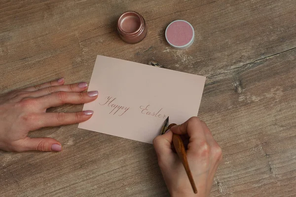 Female hands hold a pen for calligraphy. Jar with pink pearl ink on a wooden table. Easter card with lettering happy easter