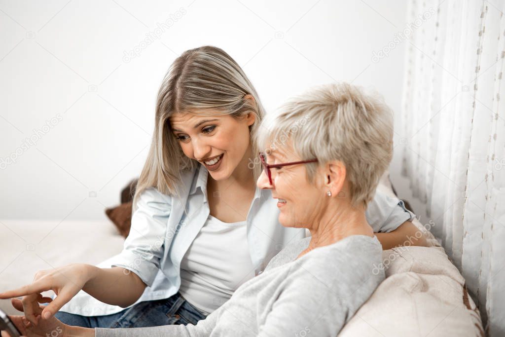 A senior blonde woman has a tablet in her hand while a young wom