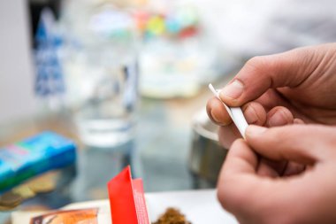 Elle tütün sigarası ve bir kağıt yaprağı yapan bir adamın eline yakın çekim. Arka planda bir kül tablası ve dökülmüş tütün görebilirsiniz. Boşluğu Kopyala.