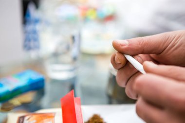 Tütün sigarasını ve bir kağıt parçasını eliyle saran bir adamın ellerine yakın plan. Arka planda bir kül tablası ve dökülmüş tütün görebilirsiniz. Boşluğu Kopyala.