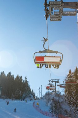 Dağda güneşli güzel bir gün, güneş altı koltuklu bir asansörle parlıyor. Teleferiğin altında kayak pisti, kayak pisti ve çam ormanı var..