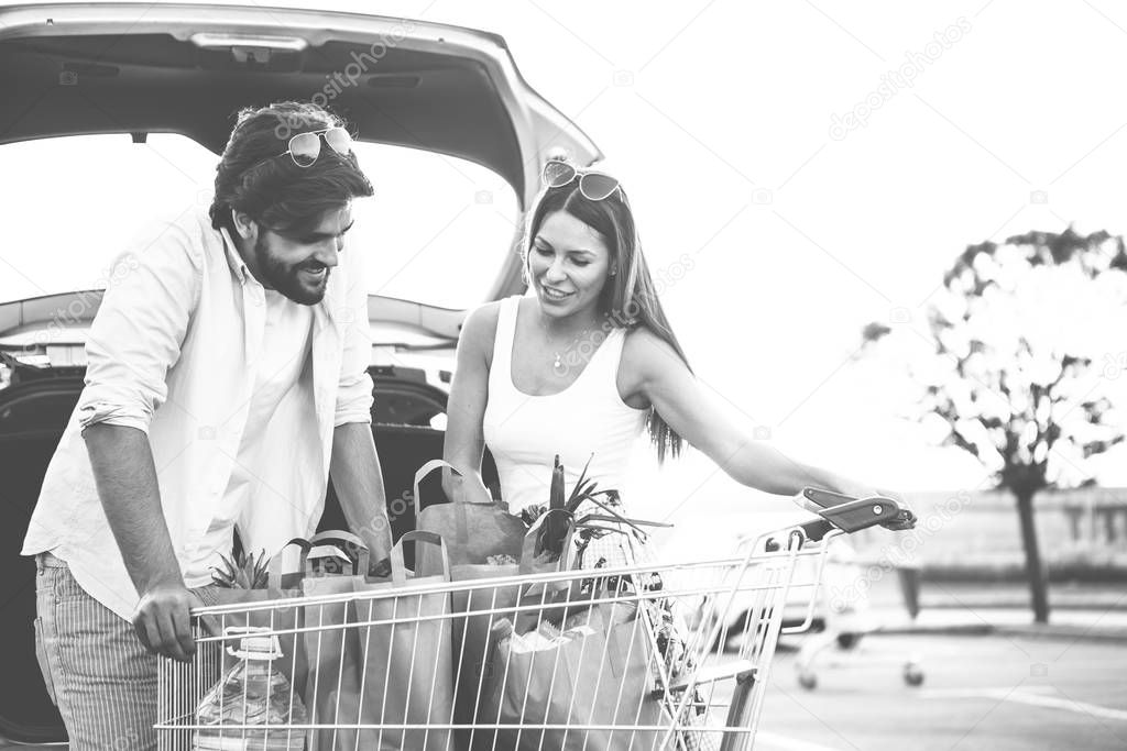 The young couple ended up buying food at a super market. They transfer the groceries from the cart to the car and make sure they have purchased everything they need. Black and white photo.