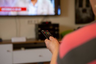 Son sınıf öğrencisi bir kadın televizyon seyrediyor. Kanalı değiştirmek istediği için televizyonu işaret ettiği uzaktan kumandayı elinde tutuyor. Odak noktası uzaktan kumanda..