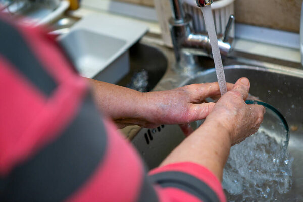 A senior woman is over the sink and she washes the glass bowl under a jet of water. The focus is on her hands.