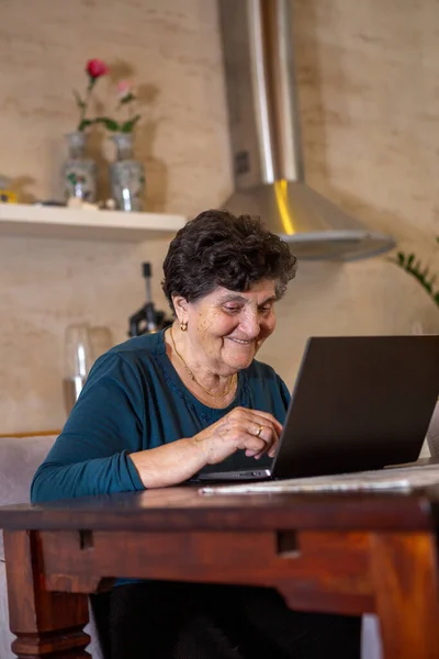 A senior woman with black short hair in a turquoise shirt is typing on a gray laptop keyboard. She laughs and is thoughtful as she looks at the monitor from her laptop computer. She is at the wooden dining table in the kitchen.