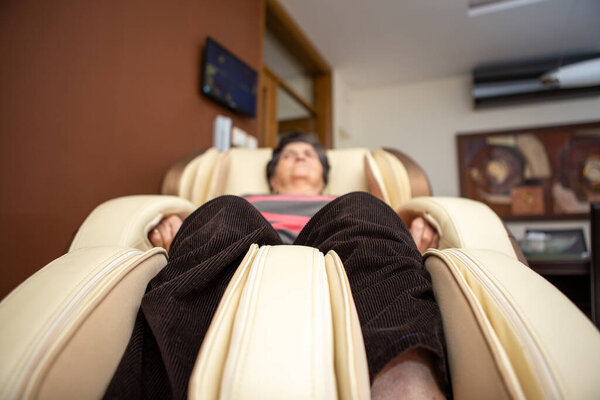A senior woman lies on a massage chair in her apartment. She really adores massaging herself in this massage chair and enjoys it. Focus on the bottom of the massage chair.
