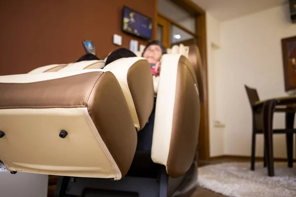 A senior woman lies on a massage chair in her apartment. She really adores massaging herself in this massage chair and enjoys it. Focus on the bottom of the massage chair.