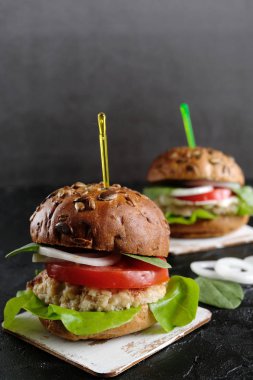 Domatesli vejetaryen hamburger, marul, soğan, fasulye pirzolası ve koyu arkaplanda tahıllı çörek. Her zamanki hamburgerin alternatifi. Sağlıklı beslenme kavramı. Dikey. Boşluğu kopyala.