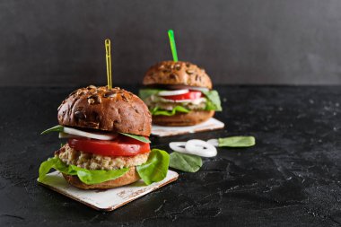 Domatesli vejetaryen hamburger, marul, soğan, fasulye pirzolası ve koyu arkaplanda tahıllı çörek. Her zamanki hamburgerin alternatifi. Sağlıklı beslenme kavramı. Boşluğu kopyala.