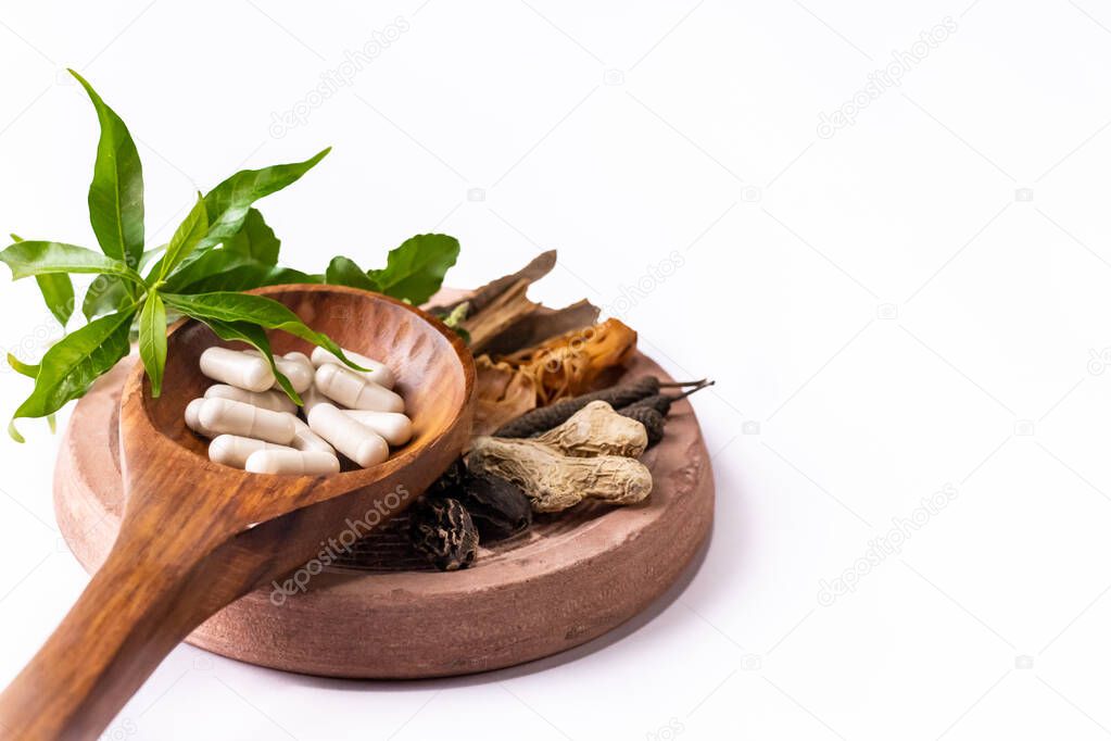 High angle close-up shot of assorted herbs and whole spices like ginger, nutmeg, aril, black pepper and tulsi with herbal capsules in a wooden spatula on white background copy space
