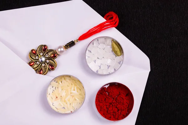 Hint Festivali - Pilav taneleri, kumkum, şeker kayası ve kakule kaplı zarif Rakhi beyaz bir zarfın içinde, siyah arka planda. Raksha Bandhan konsepti