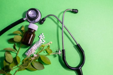 Homeopati haplarının ve steteskoplu sıvı cam şişenin üst görüntüsü ve nane rengi arka planda yapraklar.