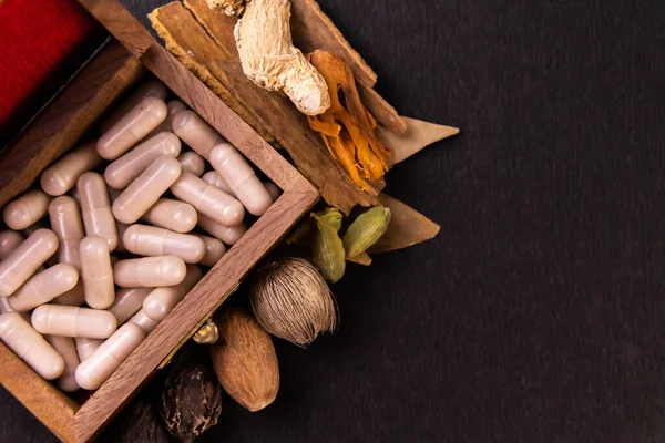 Bitkisel tıp konsepti, ahşap bir kutuda ayurvedic tabletlerin üst görüntüsü ve siyah arka planda dağınık baharatlar.