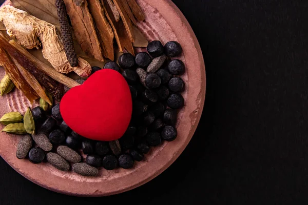 Top view of a heart shape with herbal pills and dried spices on a grinding stone over black background. Cardiovascular diseases and treatment concept