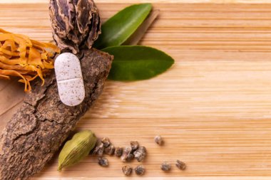 Bitkisel tıp konsepti. Yeşil yapraklı tek bir beyaz ayurvedik tabletin üst görüntüsü, tarçın çubuğu, tahtadan arka planda tohumları olan siyah ve yeşil kakule.