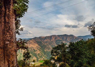 Dağların dramatik görüntüsü ve ön planda ağaç gövdesi olan bulutlu mavi gökyüzü.
