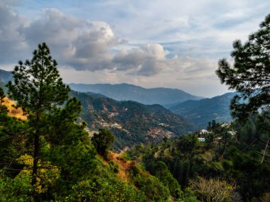 Ağaçlarla kaplı güzel dağların geniş görüntüsü ve arka planda bulutlu mavi gökyüzü.