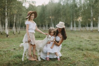 Anne ve kızları bir çiftlikte yaz günü, beyaz elbiseli bir keçiyi otlakta besler.