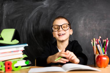 Bir okul çocuğu kameraya gülümseyen bir masada oturur, yine okulda, eğitimdedir..