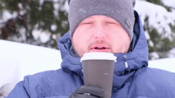 Egy szakállas férfi kávét iszik a téli sétákon, havat fúj a csészéből, havazik. — Stock videók