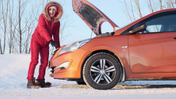 Девушка в красном зимнем красном костюме стоит рядом со сломанной машиной . — стоковое видео