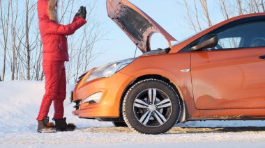A girl in a red winter red suit stands next to a broken car.
