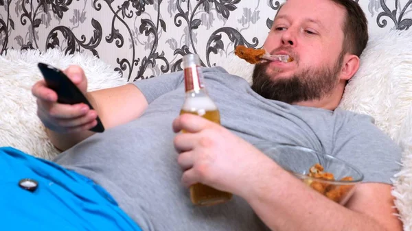 En rund, feit mann med skjegg i grå T-skjorte drikker øl og spiser fast food. Isolasjonsbegrepet under viruset og usunt kosthold – stockfoto