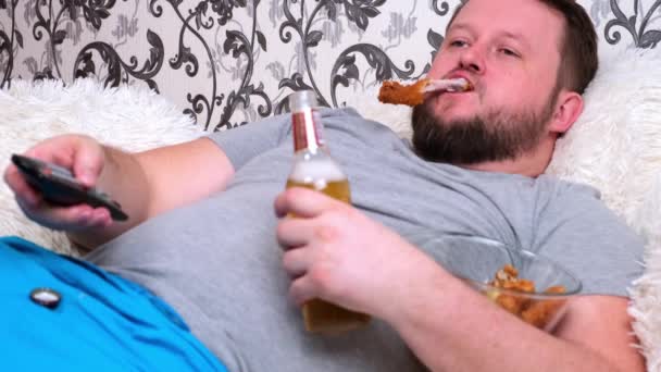 Ronde, dikke man met een baard in een grijs T-shirt drinkt bier en eet fastfood. Het concept van isolatie tijdens het virus en ongezonde voeding — Stockvideo