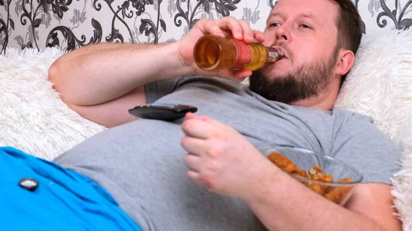 Ronde, dikke man met een baard in een grijs T-shirt drinkt bier en eet fastfood. Het concept van isolatie tijdens het virus en ongezonde voeding — Stockfoto