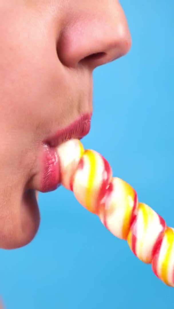 Vertical close-up, female sexy lips with red lipstick suck big spiral oblong candy lollipop. copy space. blue background — Stock Video