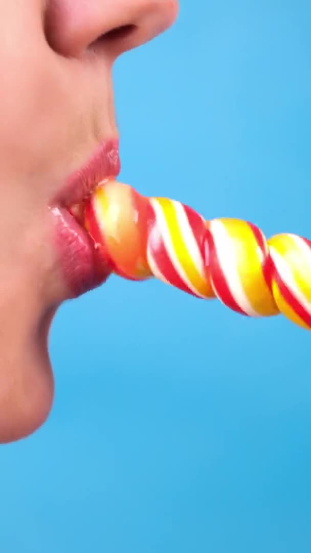 垂直クローズアップ、赤い口紅の女性のセクシーな唇は大きならせん状の楕円形のキャンディーロリポップを吸う。コピースペース。青の背景 — ストック動画