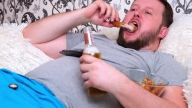 Yuvarlak suratlı, sakallı, gri tişörtlü şişman adam bira içer ve fast food yer. Virüs ve sağlıksız beslenme sırasında izolasyon kavramı