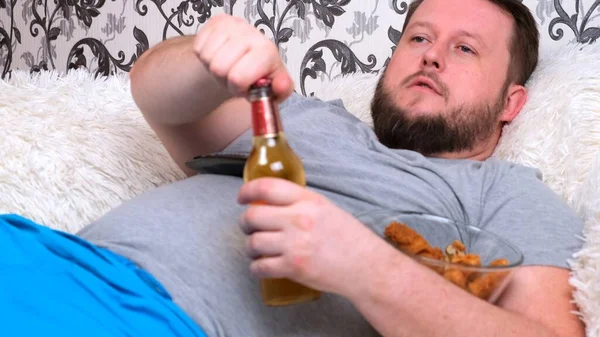 Tjukken, feit mann med skjegg i grå T-skjorte drikker øl og spiser fast food. Isolasjonsbegrepet under viruset og usunt kosthold – stockfoto