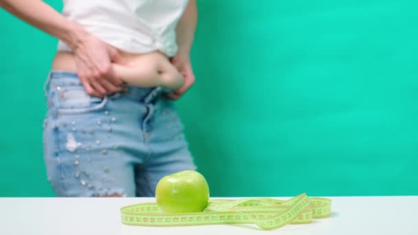Una mujer en el fondo de cinta métrica y manzana verde, toca y mide su estómago grasiento, control de peso corporal, ropa alrededor del abdomen — Vídeos de Stock