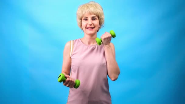 Una chica con un divertido peinado retro de 70 años hace ejercicios con pequeñas mancuernas sobre un fondo azul . — Vídeo de stock