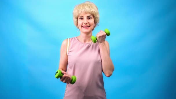 Une fille avec une coiffure rétro drôle de 70 fait des exercices avec de petits haltères sur un fond bleu . — Video