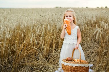 Bir kız elinde bir sepet, ekmek, çörek, un ürünleri, bir simit, mısır gevreği ekmeğiyle, yaz aylarında, gün batımında, beyaz bir elbise içinde bir buğday tarlasında duruyor.