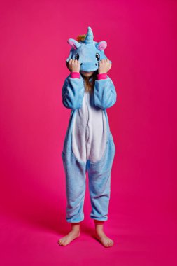 Çekici kız tek boynuzlu at kostümü içinde şakacı poz veriyor. Kigurumi 'de pembe arka planda eğlenen duygusal bir kadının stüdyo fotoğrafı..