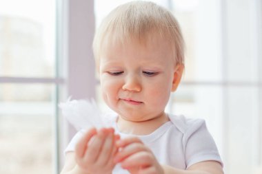 Küçük beyaz bir kız pencere kenarında tek başına oturuyor ve beyaz tüylerle oynuyor..