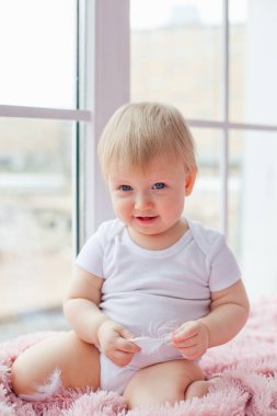 Küçük beyaz bir kız pencere kenarında tek başına oturuyor ve beyaz tüylerle oynuyor..