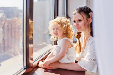 Anne ve kızı pencere eşiğinde pencereden dışarı bakıyor. Büyük bir pencerenin arkasında mutlu bir aile var. Evde kal. Ev Karantinası